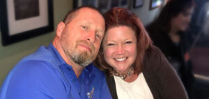 A man in a blue shirt and a woman in a white top and dark jacket smile together, embracing each other in a cozy setting with framed pictures on the wall behind them.