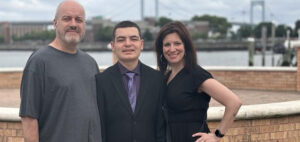 A group of three people stand outside with a waterfront and bridge in the background. A man in a gray shirt, a young man in a suit, and a woman in a black dress pose together, smiling.