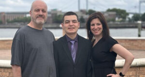 A group of three people stand outside with a waterfront and bridge in the background. A man in a gray shirt, a young man in a suit, and a woman in a black dress pose together, smiling.