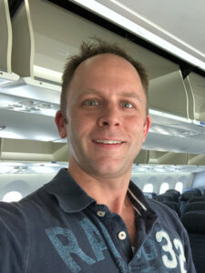 A person taking a selfie inside an airplane. They are smiling and wearing a dark polo shirt with the number 32 on it. The cabin is mostly empty, showing rows of seats and overhead compartments.