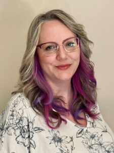 A woman with glasses, wearing a floral-patterned top, has long wavy hair with gray and purple strands. She is smiling gently against a neutral background.