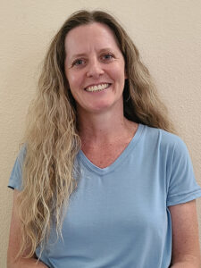 A person with long, wavy hair smiles while standing against a beige wall. They are wearing a light blue t-shirt.