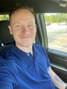 A person with short hair is smiling inside a car. They are wearing a blue polo shirt and have a tattoo on their arm. Through the window, a parking lot and some trees are visible under a clear sky.