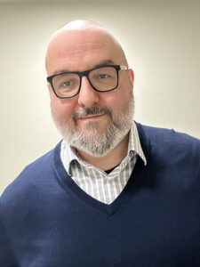 A bearded person wearing glasses, a striped shirt, and a navy blue sweater, standing against a plain light-colored background, smiling slightly.
