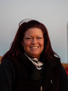 A person with long, dark red hair smiles at the camera. They are wearing a black vest and a patterned scarf around their neck. The background is softly blurred, suggesting an outdoor setting.