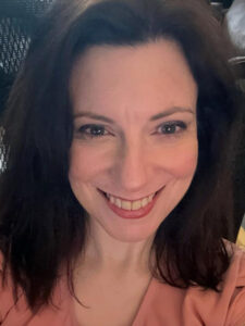 A person with dark hair is smiling at the camera. They are wearing a light peach-colored top. The background is a bit dark and blurred.