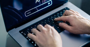 Close-up of hands typing on a laptop keyboard. The screen displays a search bar with "AI" above it, highlighting the fusion of artificial intelligence and marketing creativity. The image suggests an innovative approach where technology meets strategic imagination.