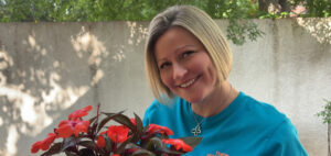 A smiling woman with short blonde hair holds a pot of vibrant red flowers. She is wearing a turquoise shirt and a silver necklace, standing outdoors in front of a light-colored wall and green trees.
