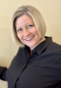 A woman with short blonde hair and a bright smile wears a black collared shirt, standing in front of a light beige wall.