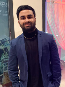 A man with a beard wearing a blue blazer over a black turtleneck and a gold chain stands indoors, smiling at the camera. There are colorful drapes and a Coca-Cola poster in the background.