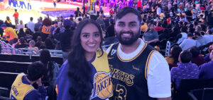 A smiling couple wearing Los Angeles Lakers jerseys pose for a photo at a crowded basketball arena, with fans and players visible in the background.