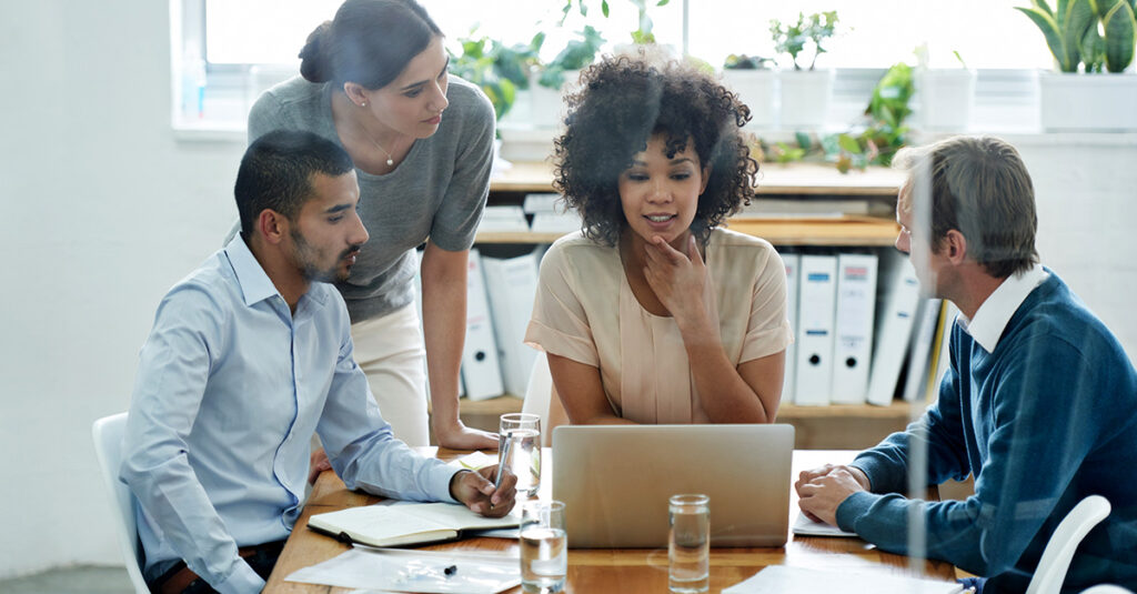 Four people are gathered around a table in an office, looking at a laptop together, discussing how The Real Risk in Telecom Isn’t Competition. It’s Silence. Papers, notebooks, and glasses of water are scattered on the table, with binders and plants in the background.