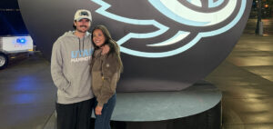 A smiling man and woman stand close together in front of a large sports team logo display at night. The ground is wet, suggesting recent rain, and the couple is dressed warmly in casual, sporty clothing.