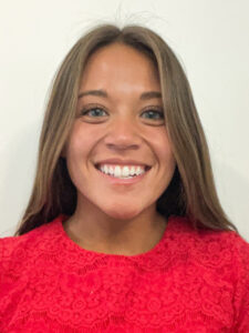 A woman with straight brown hair and green eyes smiles at the camera. She is wearing a bright red, lace-patterned top and is posed in front of a plain white background.