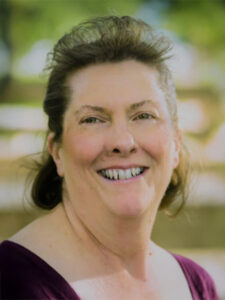 A middle-aged woman with light skin and brown hair smiles outdoors. She is wearing a dark purple top, and greenery and blurred wooden benches are visible in the background.