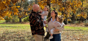A family of four stands outdoors on a sunny autumn day. The father holds one child, and the mother holds another, surrounded by trees with golden leaves. They are smiling and looking at each other.
