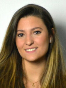 A woman with long, straight brown hair smiles at the camera. She is wearing a necklace and a dark top, with a neutral background behind her.