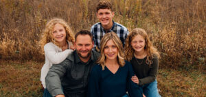 A family of five poses outdoors in a field. Two adults sit in front with three smiling children—one boy and two girls with curly hair—standing and kneeling behind them. Grassy plants and brown foliage are in the background.