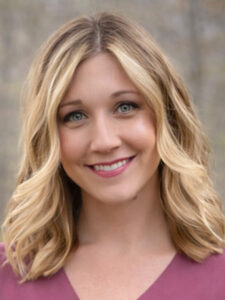 A woman with shoulder-length wavy blonde hair, light eyes, and a friendly smile wears a mauve top. The background is softly blurred, suggesting an outdoor setting.