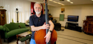 A man with gray hair and a beard wearing a black polo shirt and jeans stands indoors, resting his hands on a double bass. Elegant vintage furniture and a TV are visible in the background.