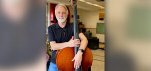 A man with gray hair and a beard, wearing a black polo shirt and jeans, stands indoors in a classroom holding an upright bass.