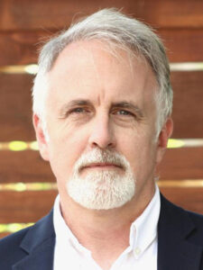 A middle-aged man with gray hair and a short white beard, wearing a white collared shirt and dark blazer, stands in front of a wooden background, looking directly at the camera with a neutral expression.