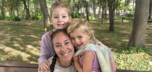 A woman with braided hair sits on a bench in a park, smiling and hugging two young girls, both smiling, with trees and greenery in the background.