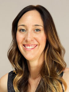 A woman with long, wavy brown hair and light highlights smiles at the camera. She is wearing a black sleeveless top and is posed against a plain, light-colored background.