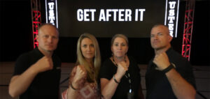 Four people stand in front of a screen displaying the words "GET AFTER IT," holding their fists up in a gesture of determination. They appear serious and focused, with a stage setup in the background.