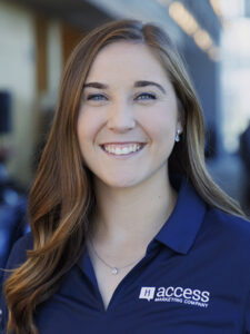 A woman with long brown hair smiles at the camera, wearing a navy blue polo shirt with the "access marketing company" logo, standing in an outdoor setting with blurred buildings in the background.