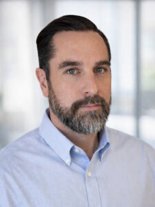 A man with short dark hair and a beard, wearing a light blue button-up shirt, stands in front of a blurred indoor background, looking at the camera with a neutral expression.