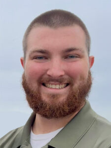 A young man with a short brown beard and buzzed hair smiles at the camera. He is wearing a green collared shirt and stands outdoors against a cloudy sky.