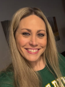 A woman with long blonde hair smiles at the camera. She is wearing a green shirt, and the background is softly lit with some blurred wall art visible.