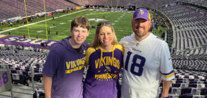 Three people wearing Minnesota Vikings apparel smile and pose together in a stadium with a football field in the background. The seats around them are mostly empty.