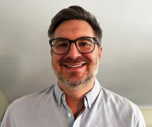 A man with short brown hair, a beard, and glasses smiles at the camera. He is wearing a light blue button-up shirt and is standing indoors against a plain light-colored wall.