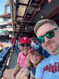 A smiling family of three wearing Phillies gear sits in blue stadium seats at a baseball game. The father wears sunglasses, the mother wears a red Phillies cap, and the child wears a navy cap and T-shirt, all enjoying a sunny day.