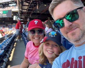 A man, woman, and child wearing Phillies gear smile for a selfie at a baseball stadium, with empty blue seats and other fans in the background.