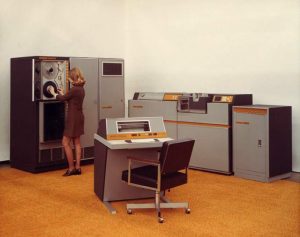 A person operates a large, vintage computer system featuring multiple consoles and cabinets in a room with an orange carpet. The setup includes tape drives and a central control panel with buttons and switches.