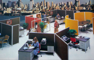 A retro office with various cubicles and typewriters, featuring employees working or interacting amidst colorful partitions. A city skyline is prominently visible through the windows, suggesting a vibrant urban setting.