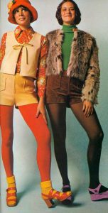 Two women in vibrant 1970s fashion. The one on the left wears a red hat, floral blouse, yellow vest, shorts, and orange tights. The woman on the right wears a green turtleneck, brown shorts, furry jacket, and dark tights, both with colorful shoes.