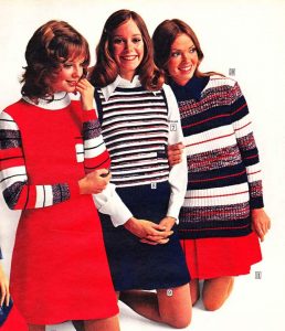 Three women, dressed in vintage 1970s outfits with knit designs and stripes, are posing confidently like marketing consultants redefining fashion. The bold colors of red, blue, and white shine through their skirts and long-sleeved sweaters as they smile warmly.