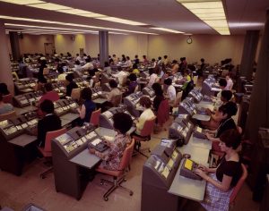 A large room filled with rows of women working at vintage mechanical desks with typewriter-like machines. The room has fluorescent lighting, and a clock is visible on the back wall. The women are dressed in mid-20th-century office attire.