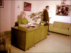 A woman with blonde hair sits at a large wooden desk, looking at papers. A man in a suit stands nearby, facing away. The room has green furniture, plants, and colorful artwork on the walls.