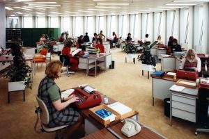 A large office space with many desks, each equipped with typewriters and paperwork. Several people are working at their desks. The room has large windows and potted plants are placed between desks. The environment looks busy and organized.
