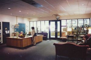 A library interior with a person standing at a reception desk while another person sits behind it. Bookshelves are visible in the background, along with a cozy seating area featuring chairs and a sofa. Natural light filters through large windows.