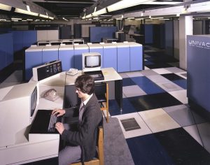 A person is working at a retro computer terminal in a large room filled with blue hardware units labeled UNIVAC. The flooring is a checkered pattern of black, white, and blue.