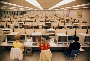 A large room filled with rows of people working at vintage computer stations. Three women in colorful clothing sit in the foreground, focused on their screens. The space has a uniform, structured layout with beige partition desks.