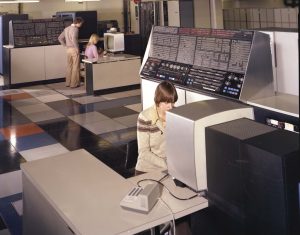 A vintage computer room with three people working. Two individuals are seated at terminals with large displays, while another stands, interacting with equipment. The room has checkered flooring and old-fashioned computer consoles.