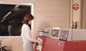 A woman in a sweater operates a large, red IBM computer in a retro office setting with beige brick walls. A fire extinguisher hangs on the wall nearby, and a window shows the room’s interior with office lighting.