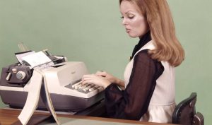 A woman with long reddish-brown hair is seated at a desk, typing on a vintage typewriter. She wears a black blouse and a sleeveless white dress. Papers are fed into the typewriter, and the background is a soft green.